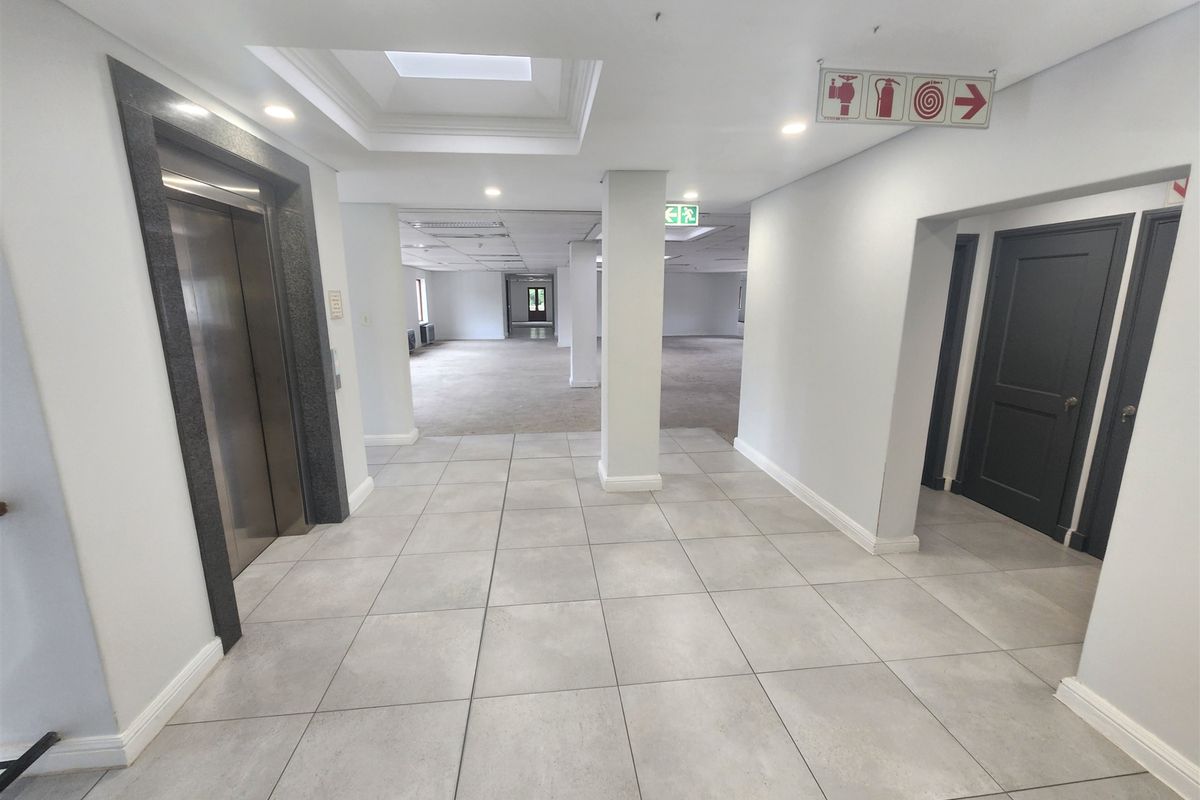 A spacious, well-lit office building hallway featuring tiled flooring, a modern elevator entrance, and clear fire safety signage.