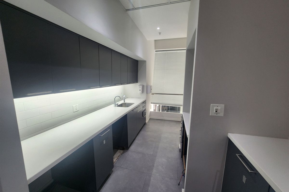Modern office kitchenette featuring dark cabinetry, white countertops, subway tile backsplash, and tiled flooring, providing a clean and functional break area.