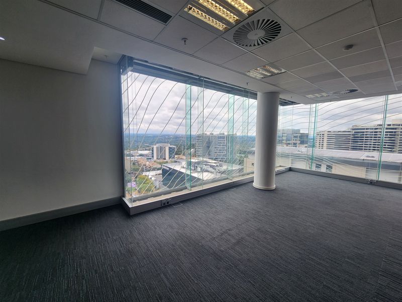 A bright, empty office space featuring floor-to-ceiling glass windows with expansive city views, carpeted flooring, and a modern suspended ceiling.