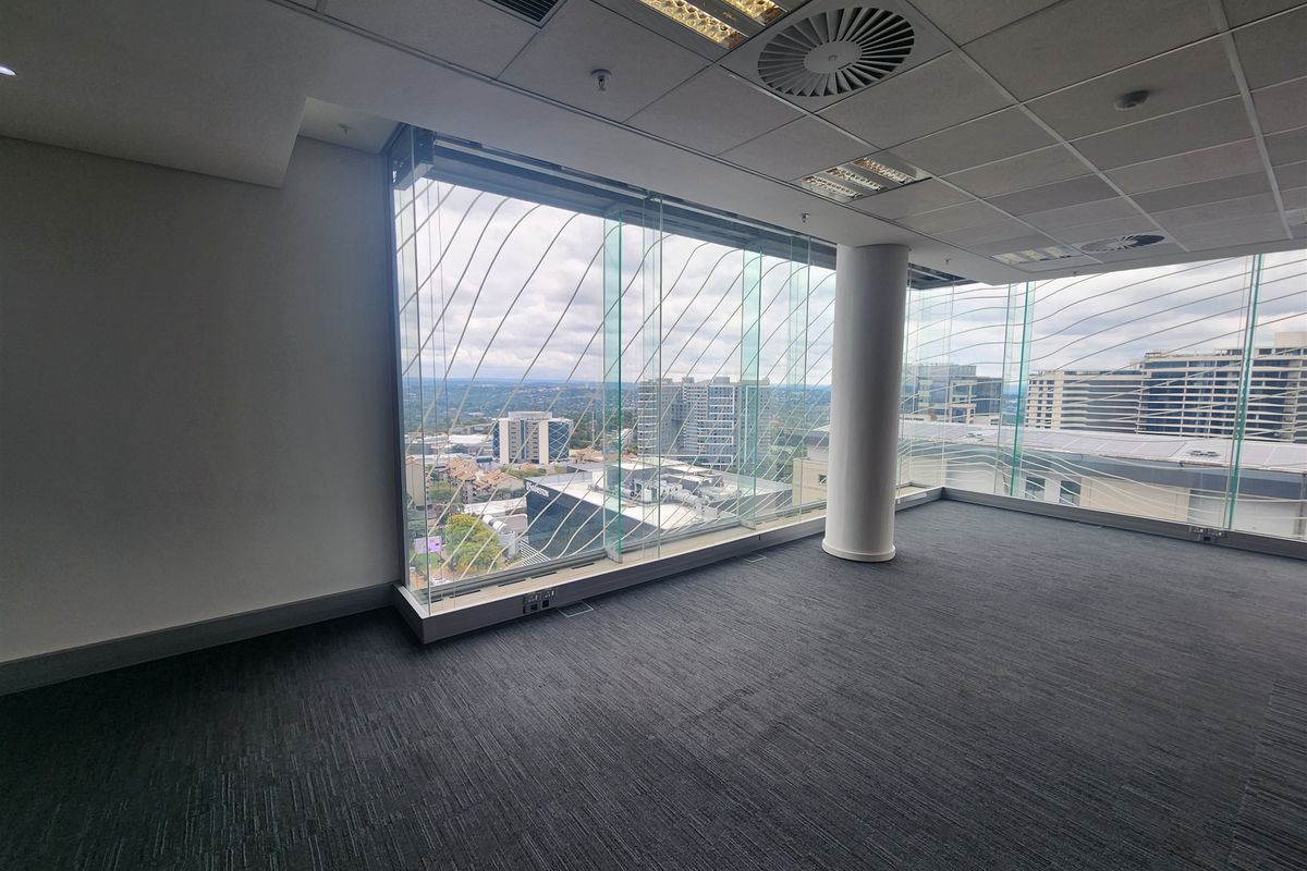 A bright, empty office space featuring floor-to-ceiling glass windows with expansive city views, carpeted flooring, and a modern suspended ceiling.