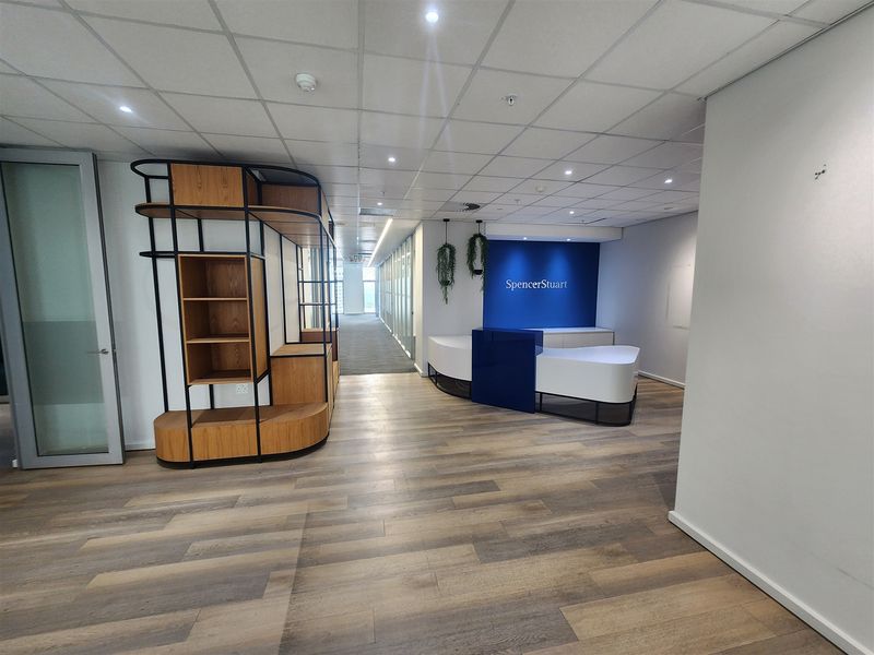 Modern office reception area featuring wood-look flooring, a built-in reception desk, suspended ceiling with recessed lighting, and glass partition walls.