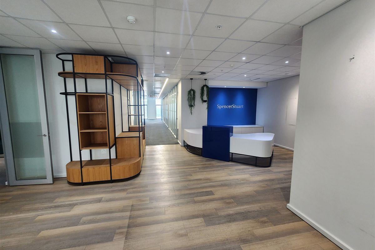 Modern office reception area featuring wood-look flooring, a built-in reception desk, suspended ceiling with recessed lighting, and glass partition walls.