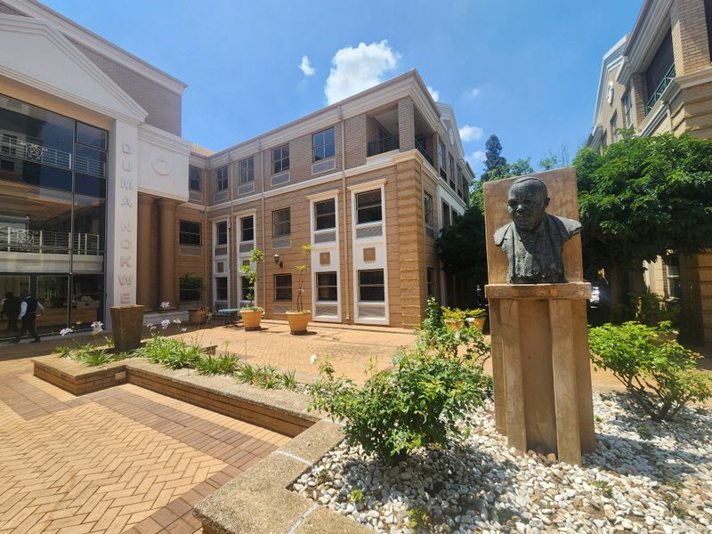 A well-maintained office building courtyard featuring brick paving, landscaped garden beds, and a prominent commemorative monument.