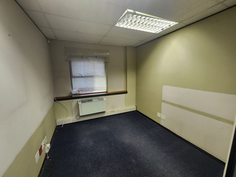 A vacant office space featuring a suspended ceiling, recessed lighting, wall-mounted HVAC unit, and carpeted flooring, requiring cosmetic updates to the walls.