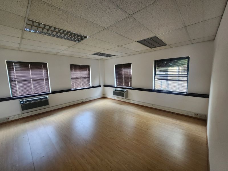 A bright, empty office space featuring laminate flooring, suspended ceilings with recessed lighting, and wall-mounted air conditioning units.