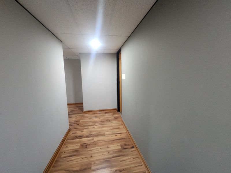 A clean, empty office hallway featuring wood-look laminate flooring, neutral grey walls, and a suspended ceiling.
