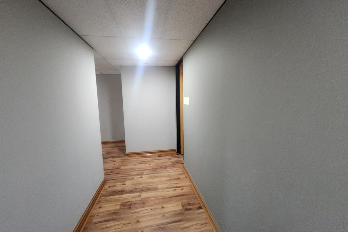 A clean, empty office hallway featuring wood-look laminate flooring, neutral grey walls, and a suspended ceiling.
