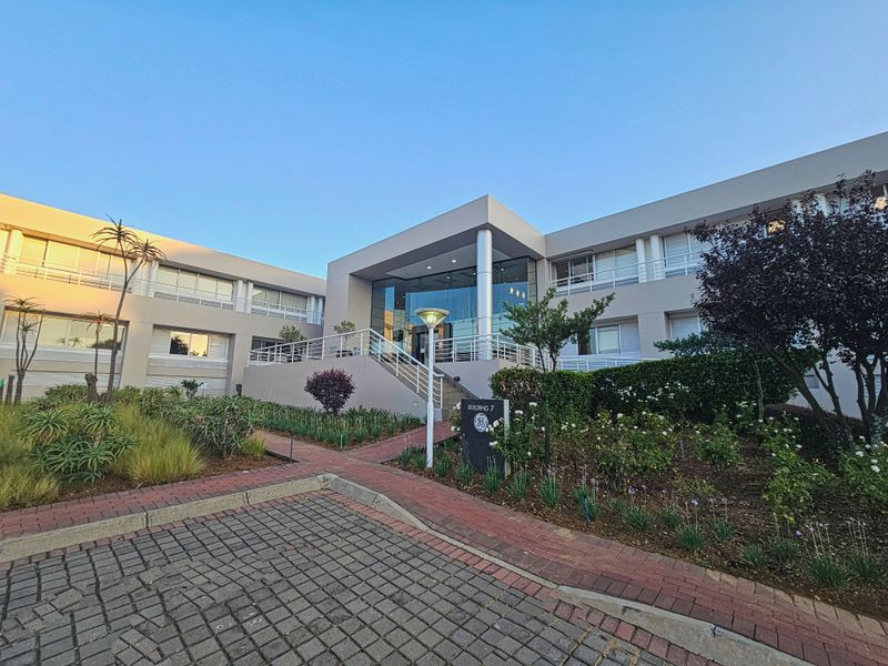 Front exterior view of a modern office building featuring a glass entrance, paved driveway, and well-maintained landscaped garden area.