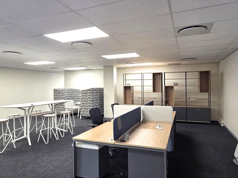 A spacious open-plan office area featuring a suspended ceiling, recessed lighting, carpeted flooring, and integrated built-in shelving units.