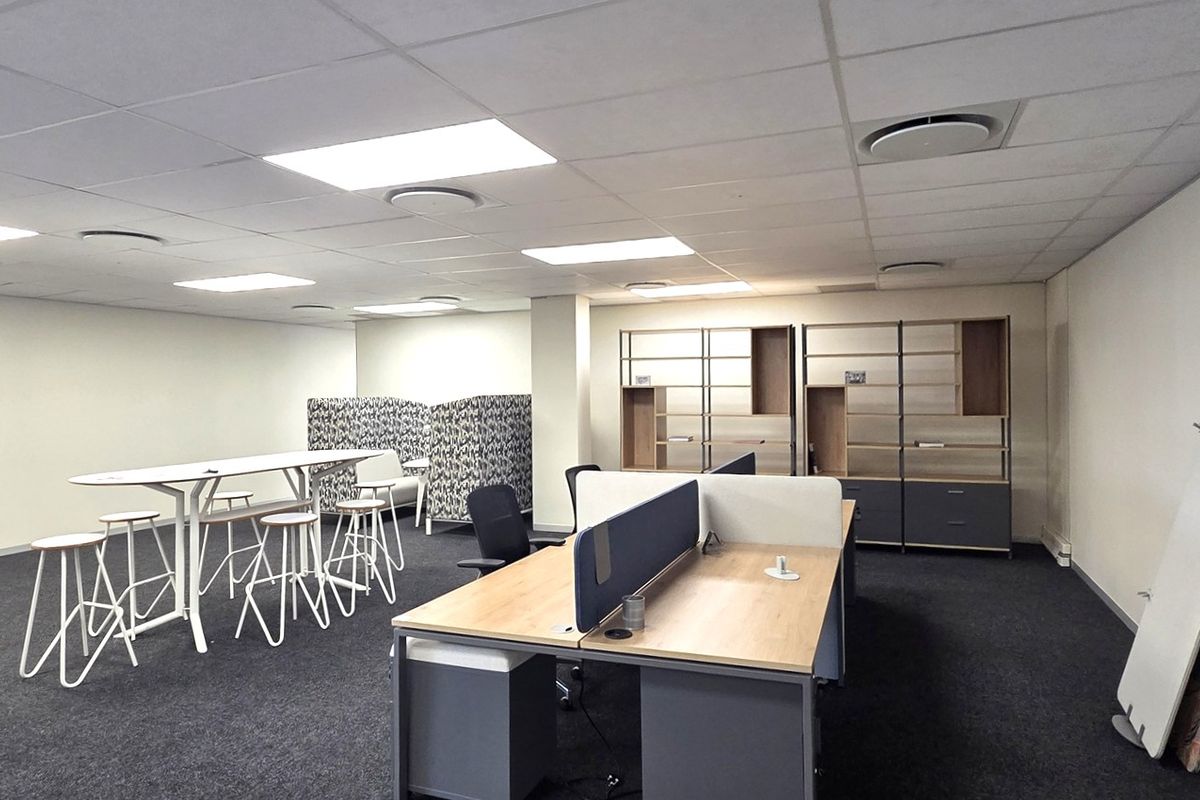 A spacious open-plan office area featuring a suspended ceiling, recessed lighting, carpeted flooring, and integrated built-in shelving units.