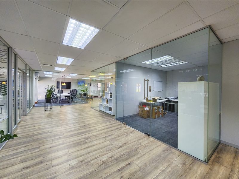 Modern office space featuring glass-partitioned private offices, suspended ceilings with recessed lighting, and a combination of wood-look laminate and carpeted flooring.