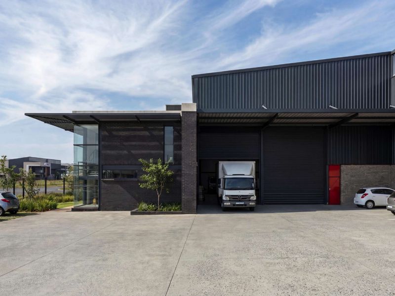 Modern industrial facility featuring a concrete driveway, metal cladding, large roller shutter doors, and a prominent glass-fronted office section.
