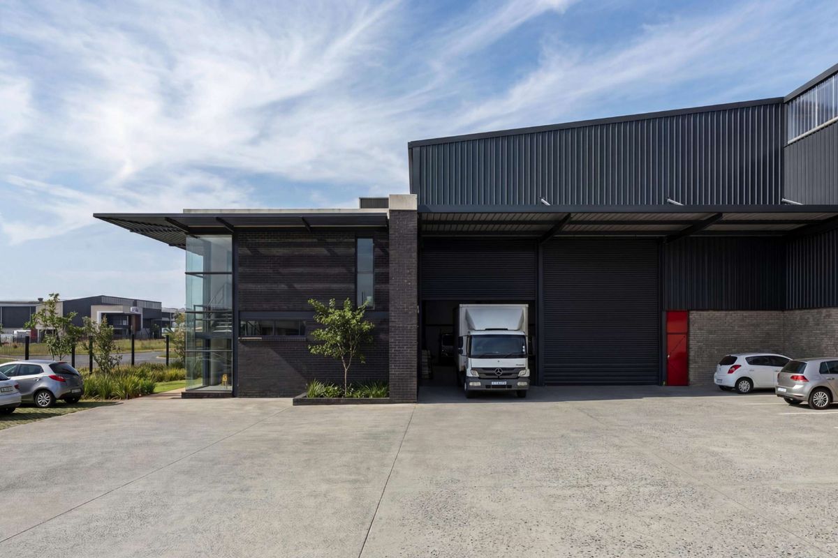 Modern industrial facility featuring a concrete driveway, metal cladding, large roller shutter doors, and a prominent glass-fronted office section.