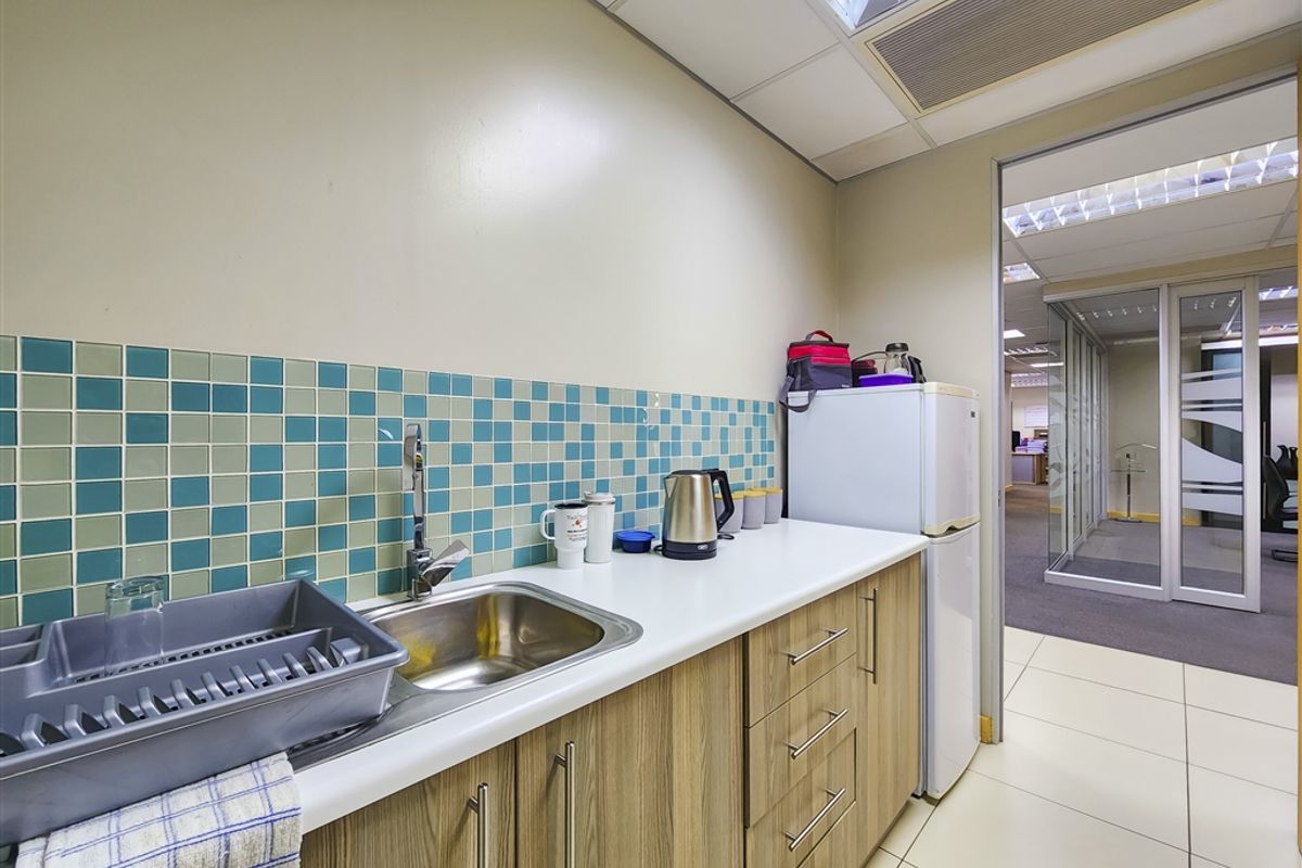 A compact office kitchenette featuring laminate cabinetry, a stainless steel sink, a vibrant tiled backsplash, and durable ceramic floor tiles.