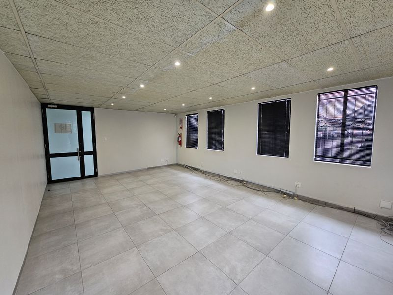 A spacious, empty office area featuring modern tiled flooring, a suspended acoustic ceiling with recessed lighting, and large windows with fitted blinds.