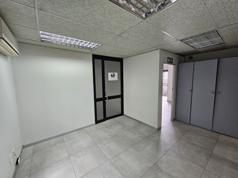 A functional office space featuring tiled flooring, a suspended ceiling with recessed lighting, built-in storage cupboards, and an air conditioning unit.