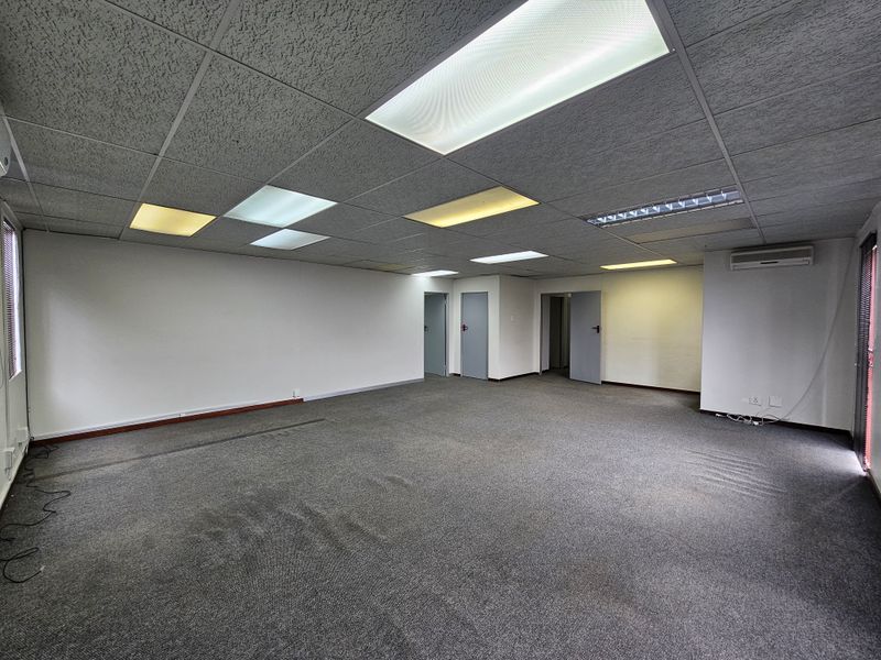 A spacious, empty office area featuring a suspended acoustic ceiling with integrated lighting, wall-mounted air conditioning unit, and grey carpeting.