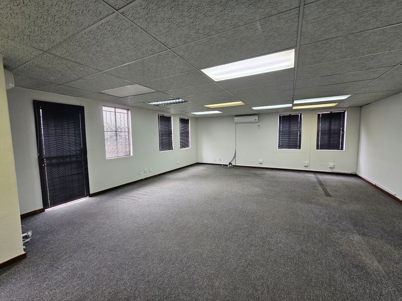 Spacious open-plan office area featuring a drop ceiling with recessed lighting, wall-mounted air conditioning unit, and large windows with fitted blinds.