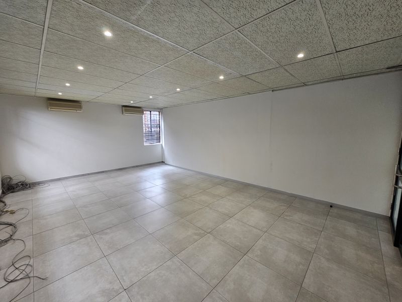 A spacious, empty office area featuring tiled flooring, a suspended acoustic ceiling with recessed lighting, and wall-mounted air conditioning units.