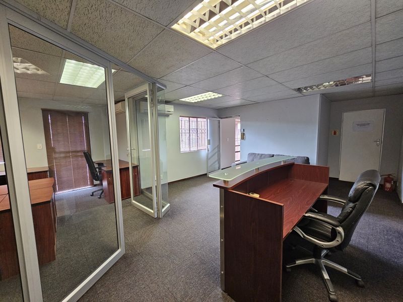 Professional office space featuring a reception desk, glass-partitioned private office, carpeted flooring, and recessed fluorescent lighting.