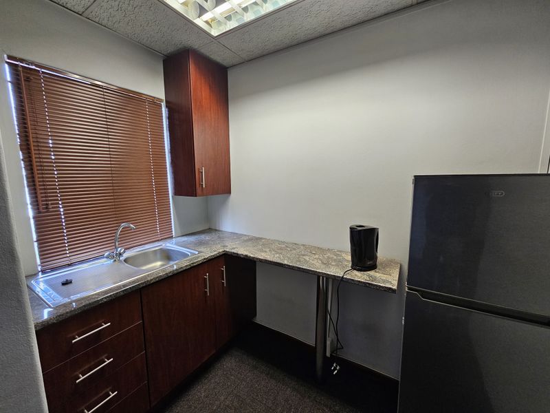 A compact office kitchenette featuring dark wood cabinetry, granite-look countertops, a stainless steel sink, and a drop ceiling with integrated lighting.
