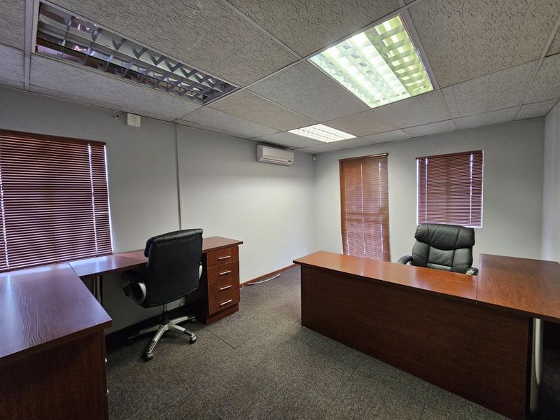 A functional office space featuring a suspended ceiling with recessed lighting, wall-mounted air conditioning unit, and fitted window blinds.