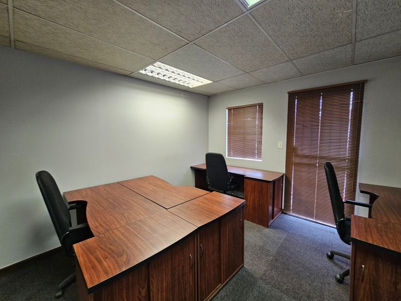 A functional office space featuring a suspended acoustic ceiling, recessed lighting, carpeted flooring, and integrated wooden desk units with window blinds.