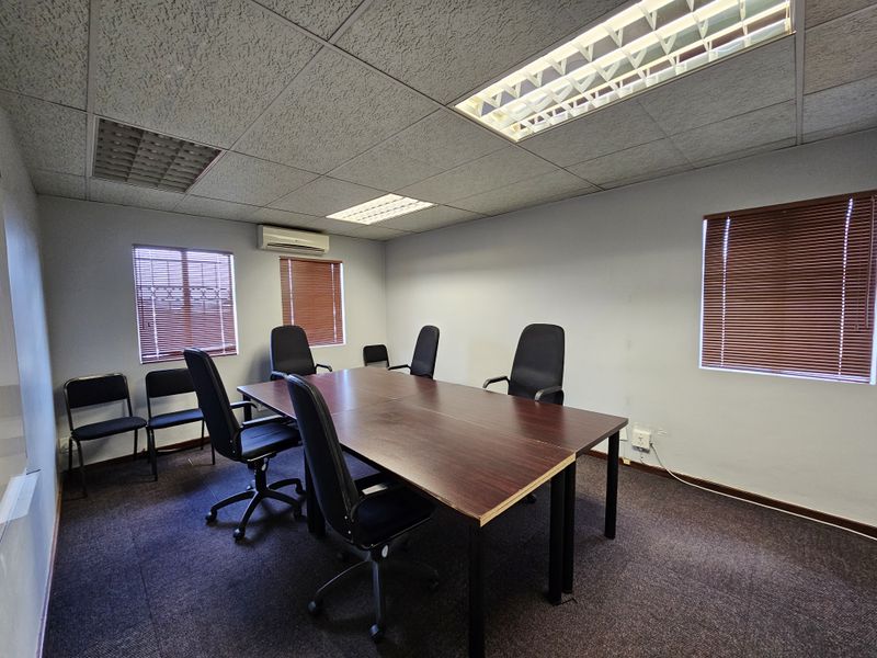 A functional office meeting room featuring a suspended ceiling with recessed lighting, wall-mounted air conditioning unit, and window blinds.