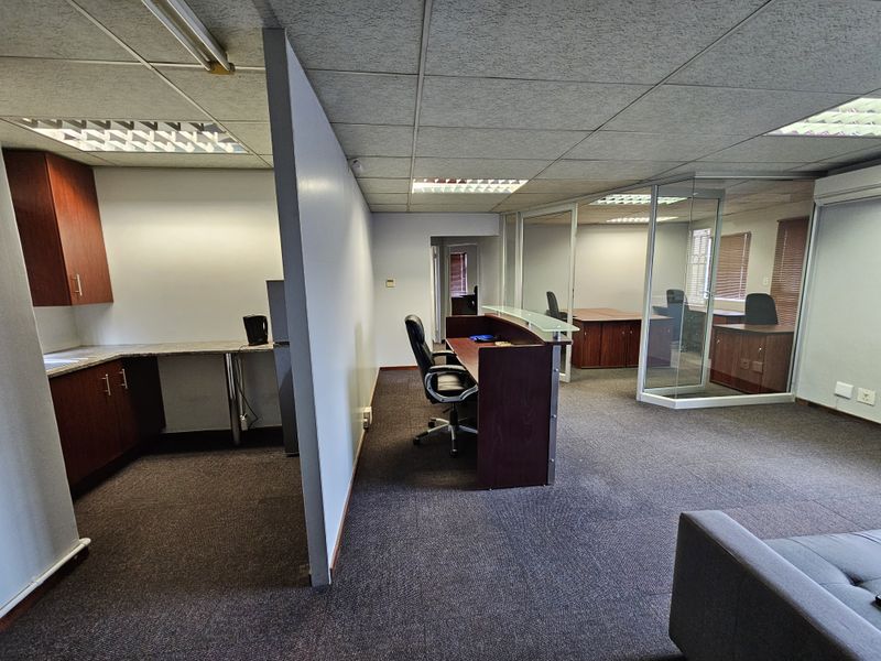 Open plan office space featuring a kitchenette area, reception desk, and glass-partitioned private office with carpeted flooring and drop ceiling lighting.