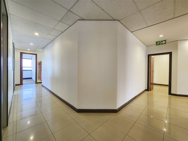 A clean, well-lit office hallway featuring tiled flooring, a suspended ceiling with recessed lighting, and clear emergency exit signage.