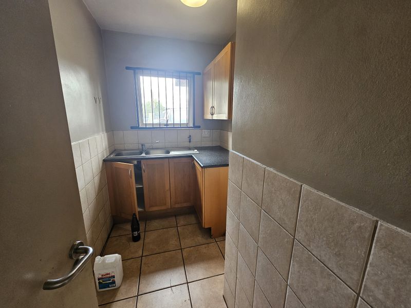 A compact scullery area featuring wooden cabinetry, a double sink, tiled splashbacks, and a security-barred window.