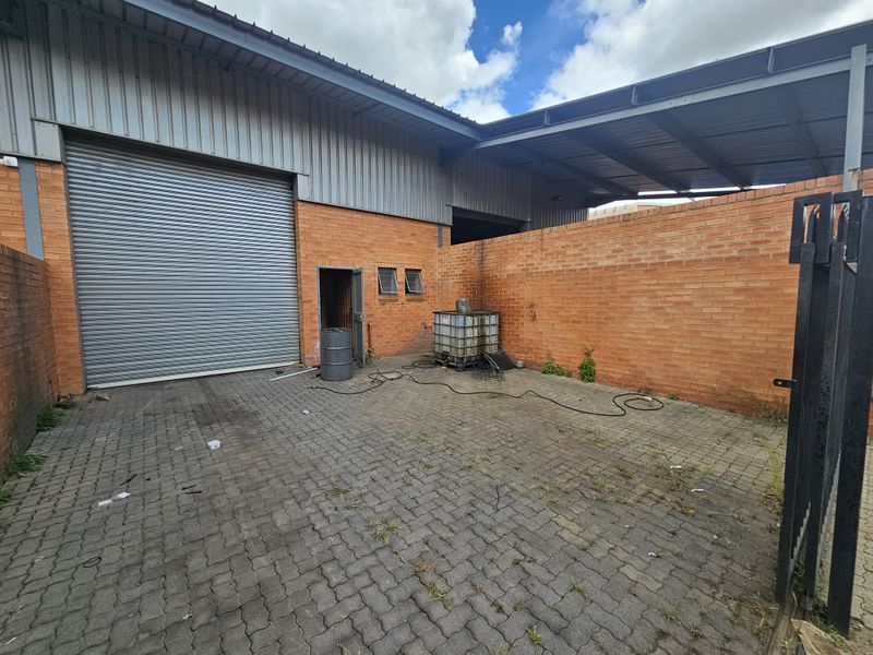Industrial exterior rear yard featuring a large roller shutter door, brick wall construction, and paved ground surface with covered storage area.