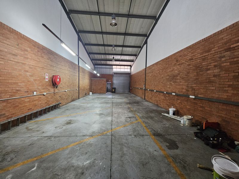 Spacious industrial warehouse unit featuring exposed brick walls, high-clearance steel truss ceiling, concrete flooring, and a large roller shutter door for easy access.