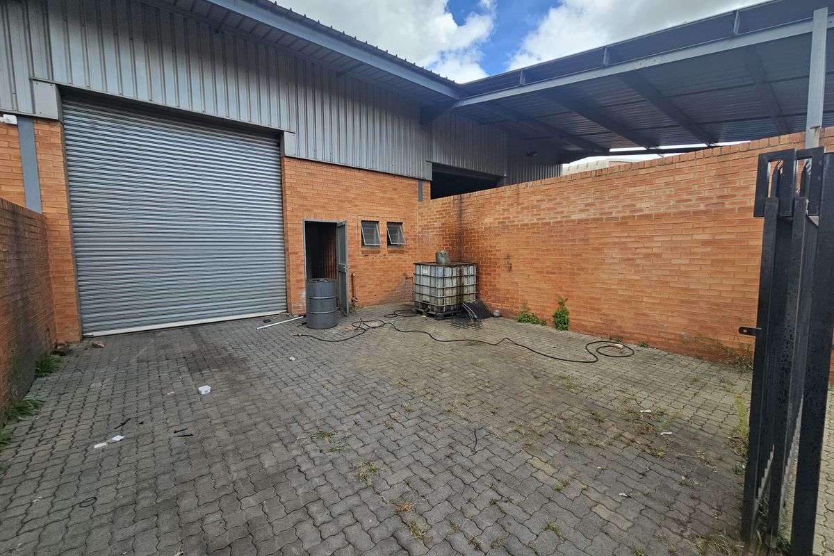 Industrial exterior rear yard featuring a large roller shutter door, brick wall construction, and paved ground surface with covered storage area.
