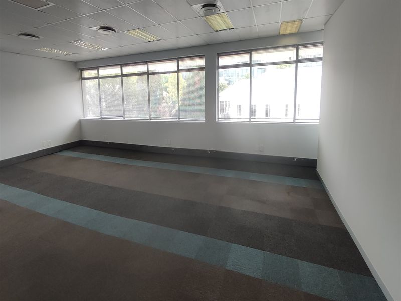 A bright, vacant office space featuring carpeted flooring, a suspended ceiling with integrated lighting, and large windows providing ample natural light.