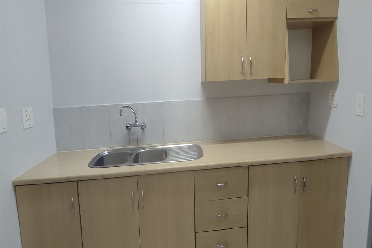 A clean and functional office kitchenette featuring light wood-tone cabinetry, a stainless steel double sink, and a neutral tiled backsplash.