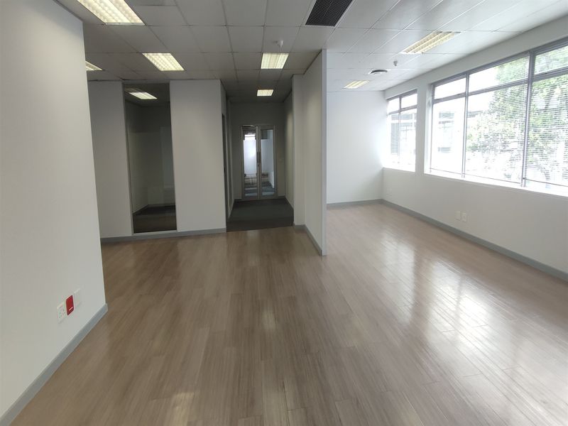 A bright, vacant open-plan office space featuring laminate flooring, large windows for natural light, and a glass-partitioned private office area.