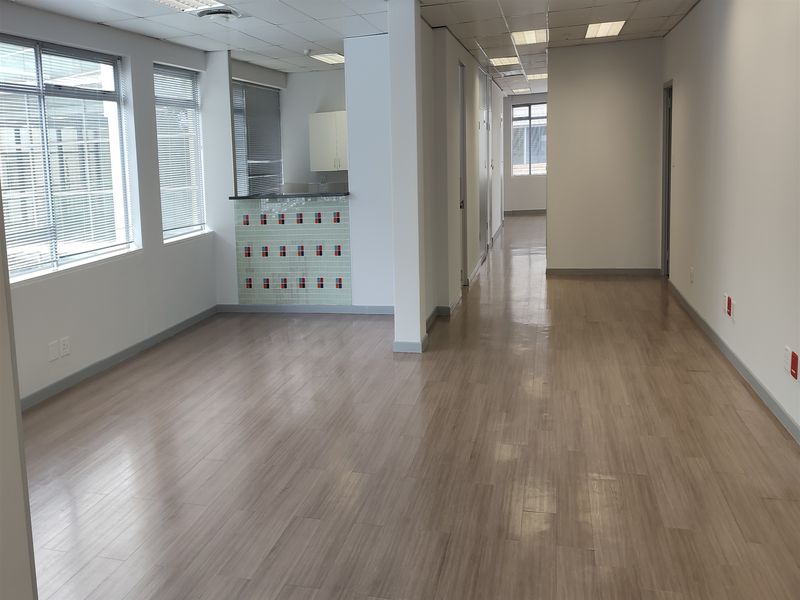 An open-plan office space featuring laminate flooring, recessed ceiling lights, large windows with venetian blinds, and a small built-in kitchenette area.