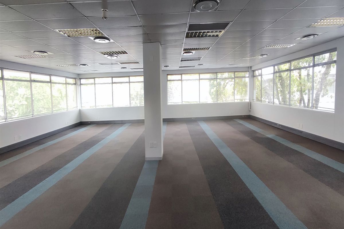 A spacious, open-plan office area featuring large windows, carpeted flooring, and a suspended ceiling with integrated lighting and air conditioning.
