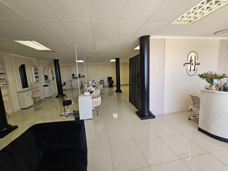 Spacious retail salon interior featuring tiled flooring, drop ceiling with recessed lighting, structural columns, and built-in reception and workstation cabinetry.