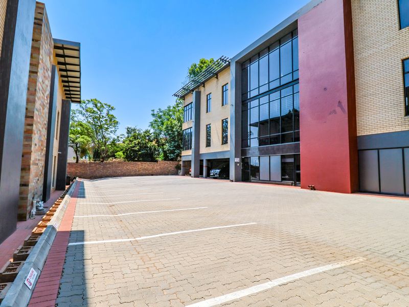 A modern office building exterior featuring a paved parking area, brick and glass facade, and covered parking bays.