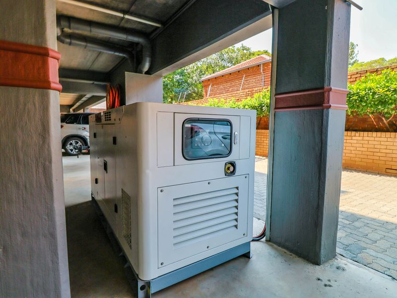 A well-maintained backup generator unit installed within a covered utility area, featuring concrete structural supports and exposed overhead piping.