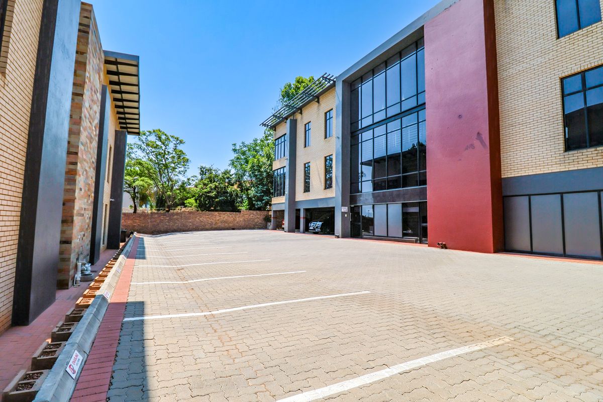 A modern office building exterior featuring a paved parking area, brick and glass facade, and covered parking bays.