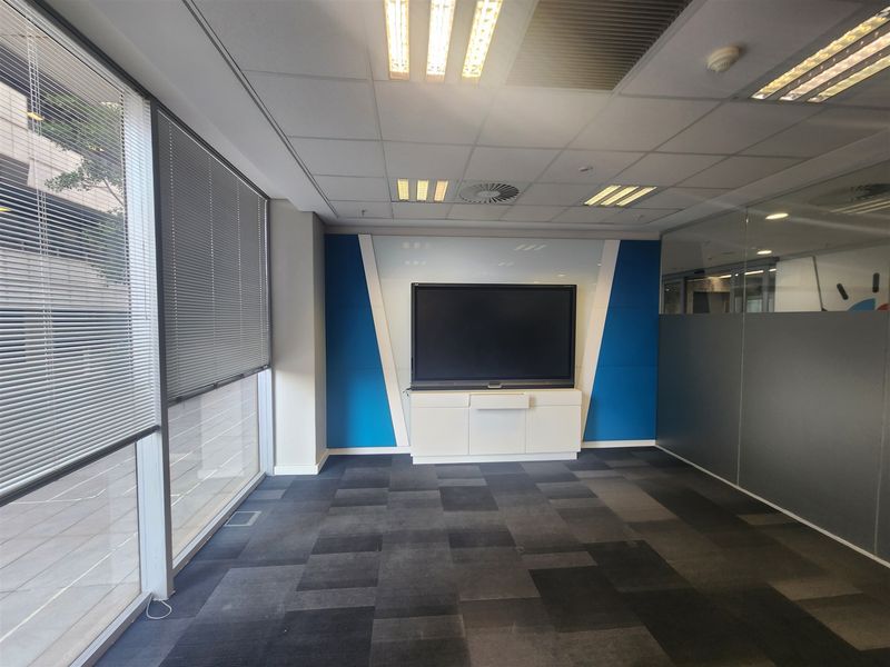 A modern, empty office space featuring carpet tiles, a glass partition wall, recessed ceiling lighting, and a built-in media console.
