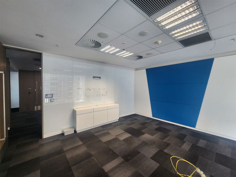 A vacant office space featuring carpet tiles, built-in cabinetry, a glass whiteboard, and a suspended ceiling with integrated lighting and air conditioning.