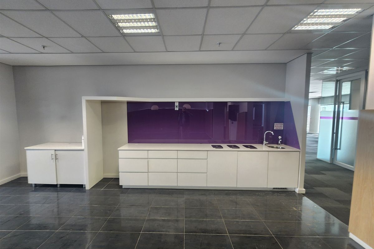 A clean, modern office kitchenette featuring white built-in cabinetry, a purple glass splashback, a sink, and dark tiled flooring.