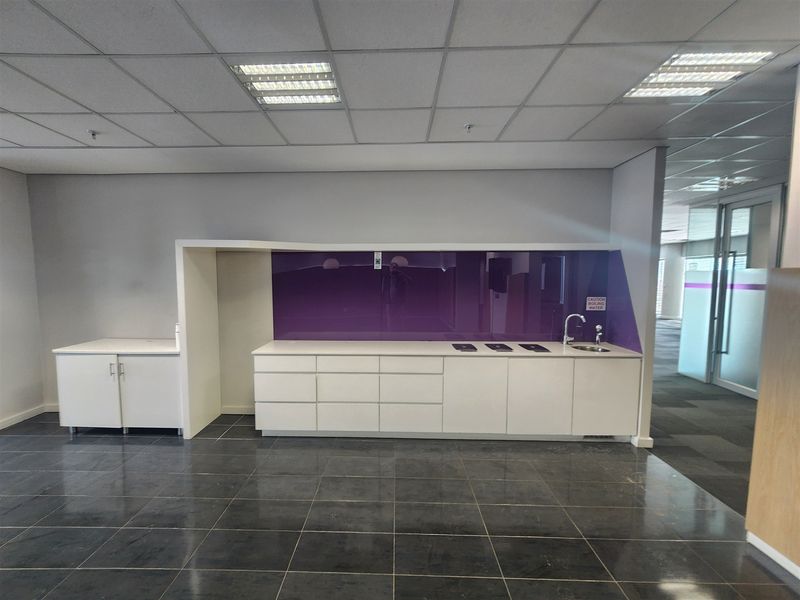 A clean, modern office kitchenette featuring white built-in cabinetry, a purple glass splashback, a sink, and dark tiled flooring.