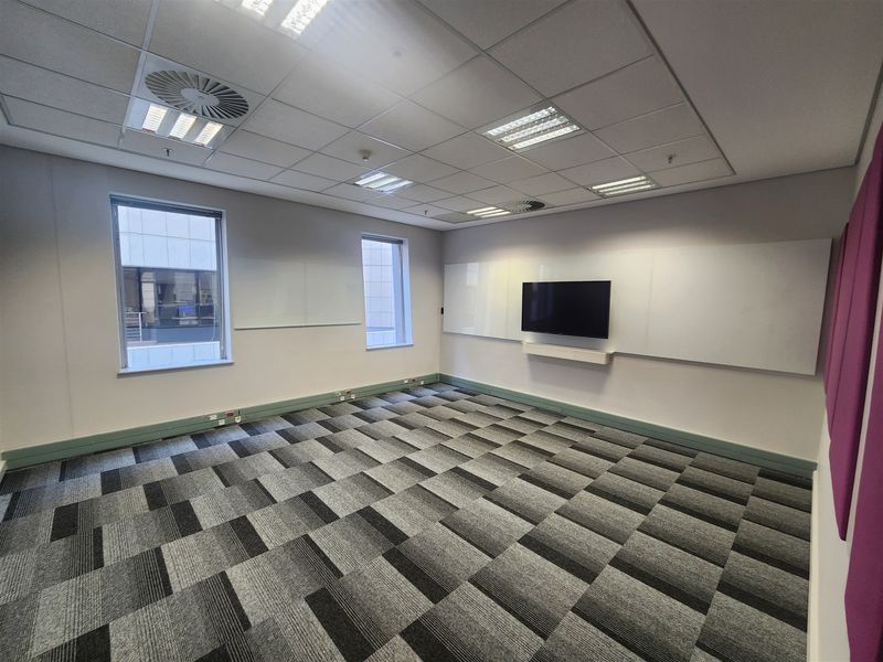 A clean, empty office space featuring carpet tiles, a suspended ceiling with recessed lighting, and wall-mounted presentation boards with an integrated television.