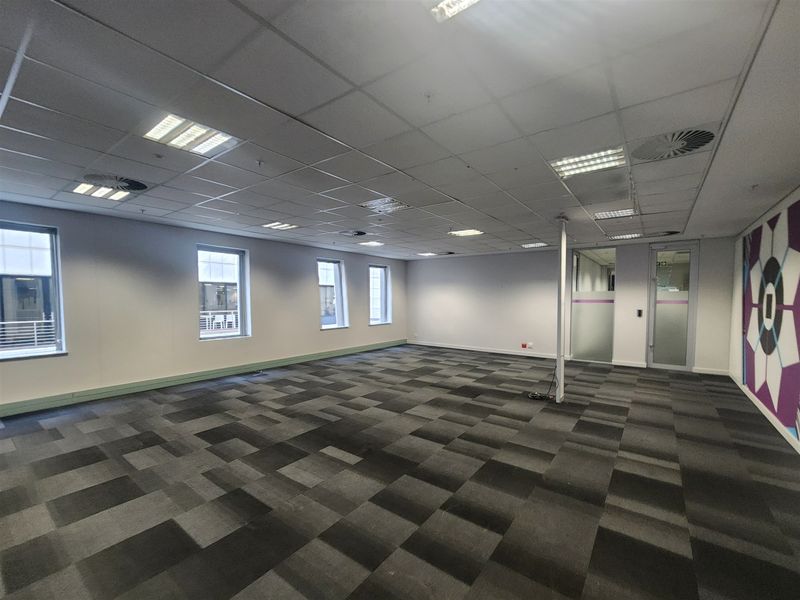 A spacious, vacant open-plan office area featuring carpet tiles, a suspended ceiling with recessed lighting, and glass partition doors.