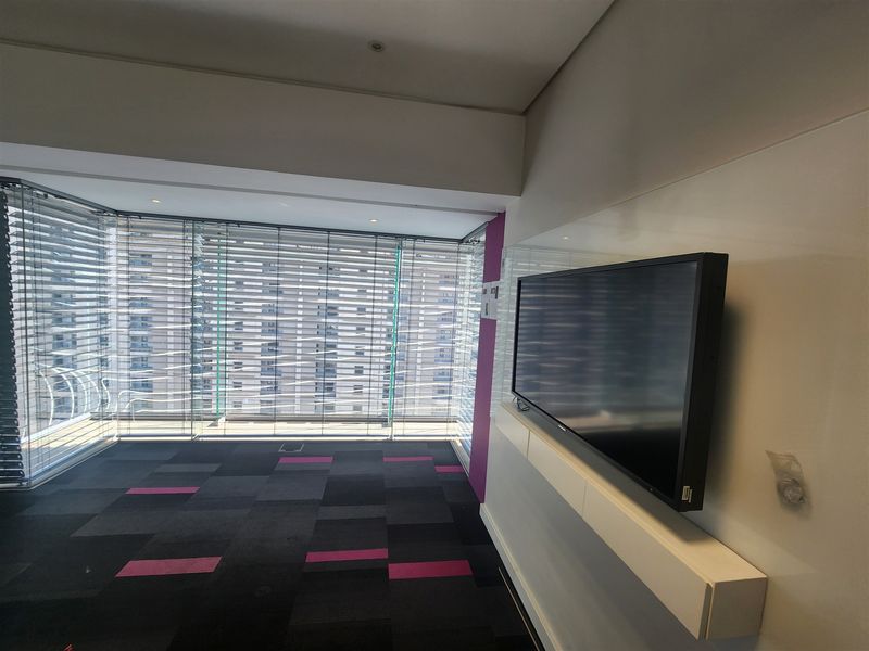 Modern office space featuring floor-to-ceiling windows with venetian blinds, patterned carpet tiles, and a sleek wall-mounted media console.